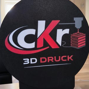 May include: A circular black sign featuring the white and red text cKr and the words 3D DRUCK in white and red. To the right of the text is a graphic of a 3D printer nozzle extruding a red layered cube. The sign is mounted on a black base.