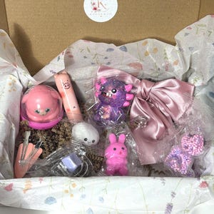 Puede incluir: Una caja de regalo de cart&oacute;n que contiene varios objetos peque&ntilde;os, incluyendo un recipiente rosa con forma de cerdo, un b&aacute;lsamo labial de cerdo, una figura de monstruo morada con purpurina, un lazo de sat&eacute;n rosa empolvado, un peluche blanco y un conejo rosa.