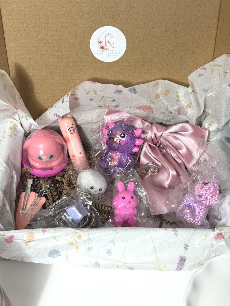 Puede incluir: Una caja de regalo de cart&oacute;n que contiene varios objetos peque&ntilde;os, incluyendo un recipiente rosa con forma de cerdo, un b&aacute;lsamo labial de cerdo, una figura de monstruo morada con purpurina, un lazo de sat&eacute;n rosa empolvado, un peluche blanco y un conejo rosa.