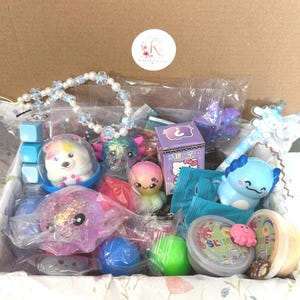 Puede incluir: Una caja de cart&oacute;n llena de varios juguetes y accesorios, incluyendo un collar de cuentas, una figura de ajolote azul, una caja misteriosa morada con Hello Kitty, un recipiente de slime, peque&ntilde;as criaturas de pl&aacute;stico y un bol&iacute;grafo azul.