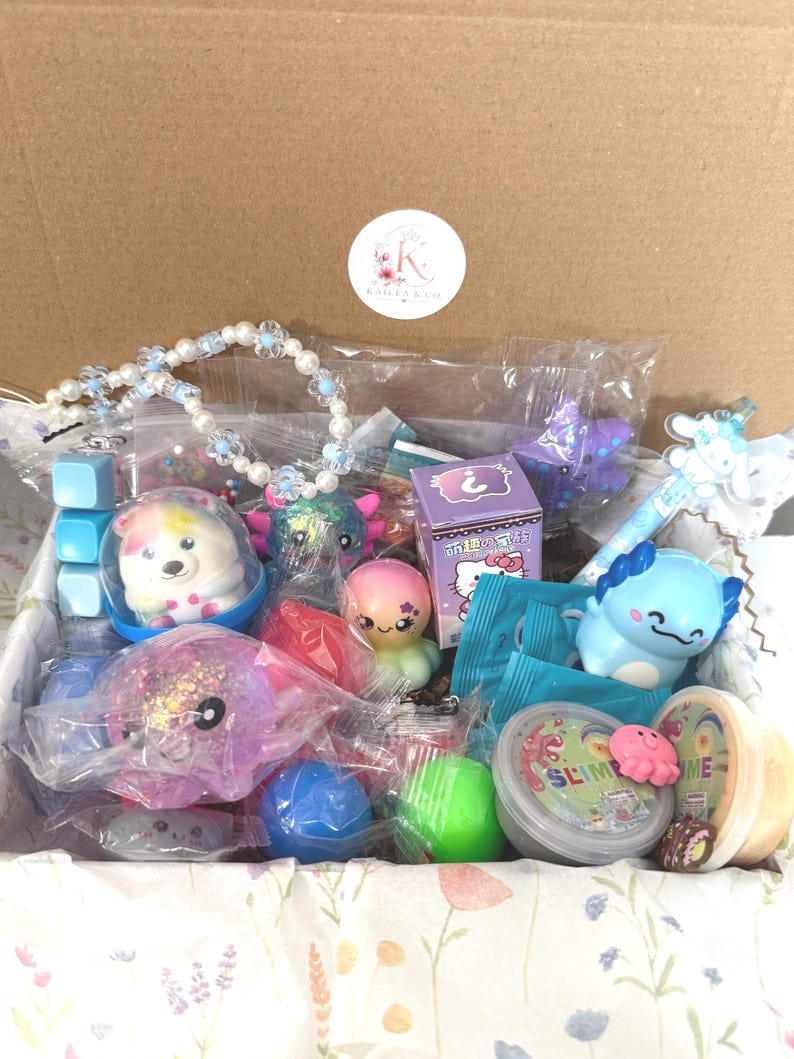 Puede incluir: Una caja de cart&oacute;n llena de varios juguetes y accesorios, incluyendo un collar de cuentas, una figura de ajolote azul, una caja misteriosa morada con Hello Kitty, un recipiente de slime, peque&ntilde;as criaturas de pl&aacute;stico y un bol&iacute;grafo azul.