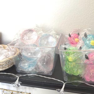 Puede incluir: Dos recipientes rectangulares de pl&aacute;stico transparente con objetos peque&ntilde;os. El recipiente izquierdo contiene esferas transl&uacute;cidas, mientras que el derecho contiene figuras de animales brillantes en rosa, azul y verde. Una cesta tejida con tapas transparentes se encuentra a la izquierda.
