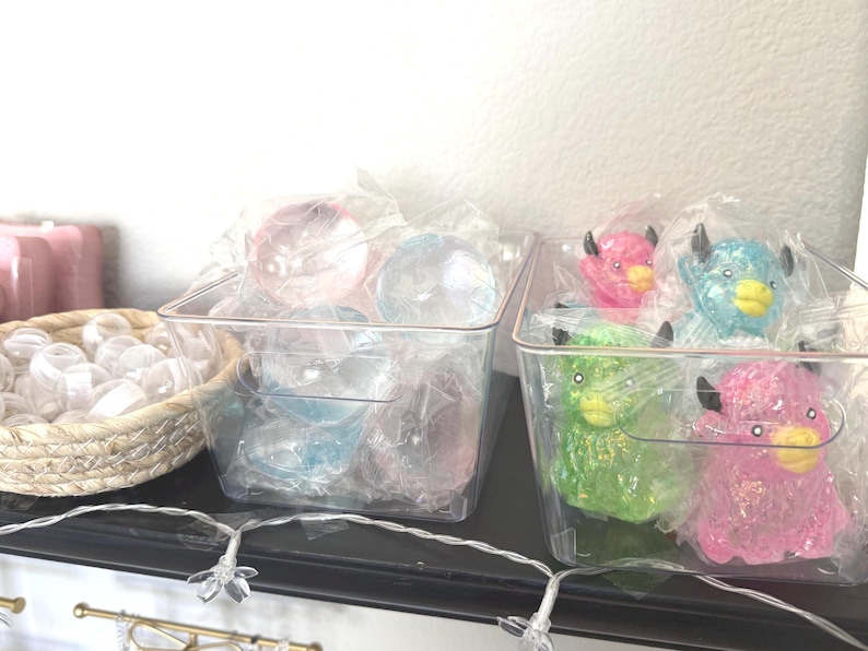 Puede incluir: Dos recipientes rectangulares de pl&aacute;stico transparente con objetos peque&ntilde;os. El recipiente izquierdo contiene esferas transl&uacute;cidas, mientras que el derecho contiene figuras de animales brillantes en rosa, azul y verde. Una cesta tejida con tapas transparentes se encuentra a la izquierda.