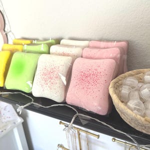 Puede incluir: Una colecci&oacute;n de bombas de ba&ntilde;o cuadradas envueltas individualmente en amarillo, verde claro, blanco y rosa, algunas con un acabado moteado. Una cesta tejida llena de peque&ntilde;os recipientes de pl&aacute;stico esf&eacute;ricos transparentes se sit&uacute;a a la derecha.