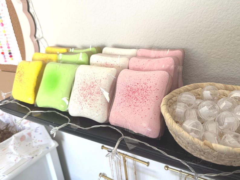 Puede incluir: Una colecci&oacute;n de bombas de ba&ntilde;o cuadradas envueltas individualmente en amarillo, verde claro, blanco y rosa, algunas con un acabado moteado. Una cesta tejida llena de peque&ntilde;os recipientes de pl&aacute;stico esf&eacute;ricos transparentes se sit&uacute;a a la derecha.