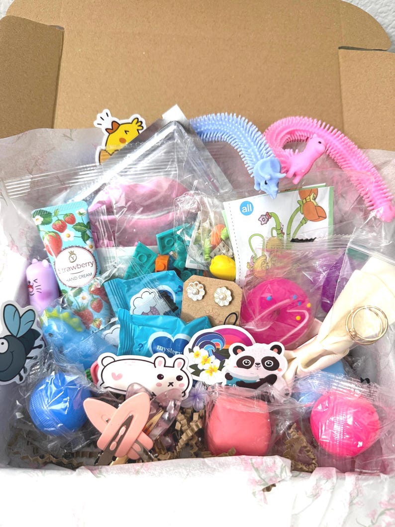 Puede incluir: Una caja de cart&oacute;n llena de art&iacute;culos peque&ntilde;os variados, incluyendo un tubo de crema de manos de fresa, un objeto rosa con forma de d&oacute;nut, un par de pendientes de flores, una pegatina de panda, un juguete de muelle azul y rosa, horquillas y abalorios.