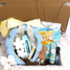 Puede incluir: Una caja de regalo que contiene una diadema de felpa azul claro con un lazo blanco, dos collares en tonos oro y plata, una bolsa a rayas amarillas, dos pinzas para el pelo azul claro, b&aacute;lsamo labial de lim&oacute;n, crema de manos y pendientes.