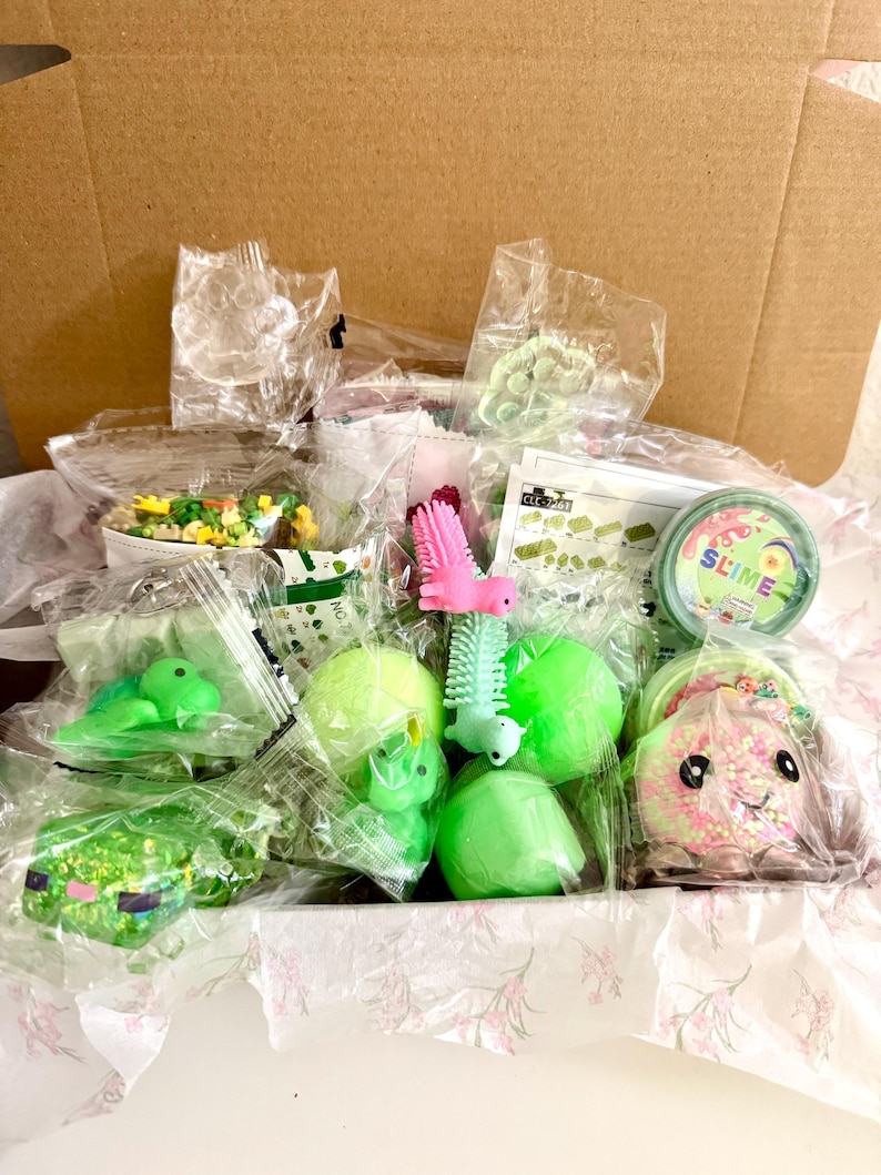 Puede incluir: Una caja de cart&oacute;n llena de varios juguetes peque&ntilde;os y art&iacute;culos sensoriales en envases de pl&aacute;stico individuales, incluyendo recipientes de slime verde, una criatura rosa con p&uacute;as, una figura de tortuga verde y un peque&ntilde;o set de construcci&oacute;n.
