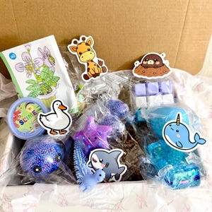 Puede incluir: Una caja de cart&oacute;n llena de varios juguetes peque&ntilde;os y art&iacute;culos en envases de pl&aacute;stico transparente, incluyendo un recipiente etiquetado SLIME, una estrella de mar morada, un narval azul, un tibur&oacute;n, un pato, una jirafa, un topo y un peque&ntilde;o set de construcci&oacute;n.