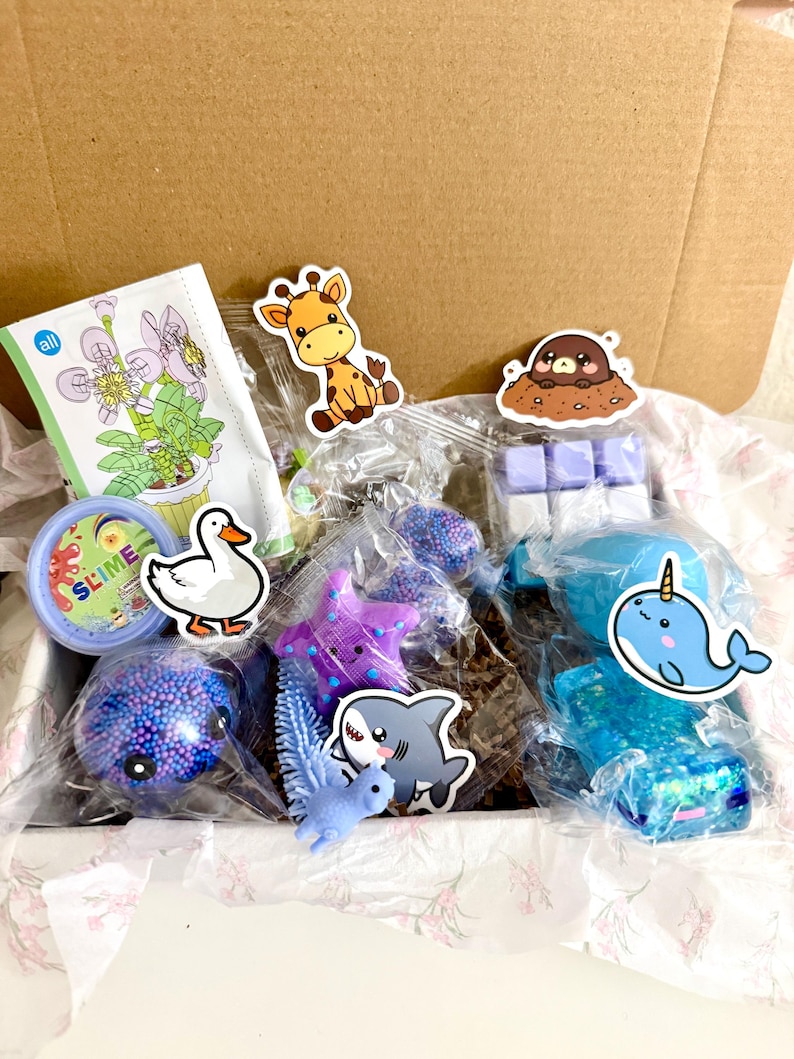 Puede incluir: Una caja de cart&oacute;n llena de varios juguetes peque&ntilde;os y art&iacute;culos en envases de pl&aacute;stico transparente, incluyendo un recipiente etiquetado SLIME, una estrella de mar morada, un narval azul, un tibur&oacute;n, un pato, una jirafa, un topo y un peque&ntilde;o set de construcci&oacute;n.
