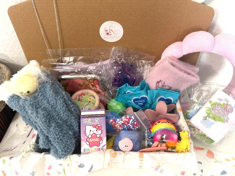 Puede incluir: Una caja de regalo de cart&oacute;n llena de art&iacute;culos variados, incluyendo un peluche azul difuso, una caja de Hello Kitty con el texto 'Luck', un peque&ntilde;o recipiente etiquetado como 'Slime', dos paquetes azules 'mystery bag', un accesorio para el pelo rosa y una peque&ntilde;a tarjeta floral.