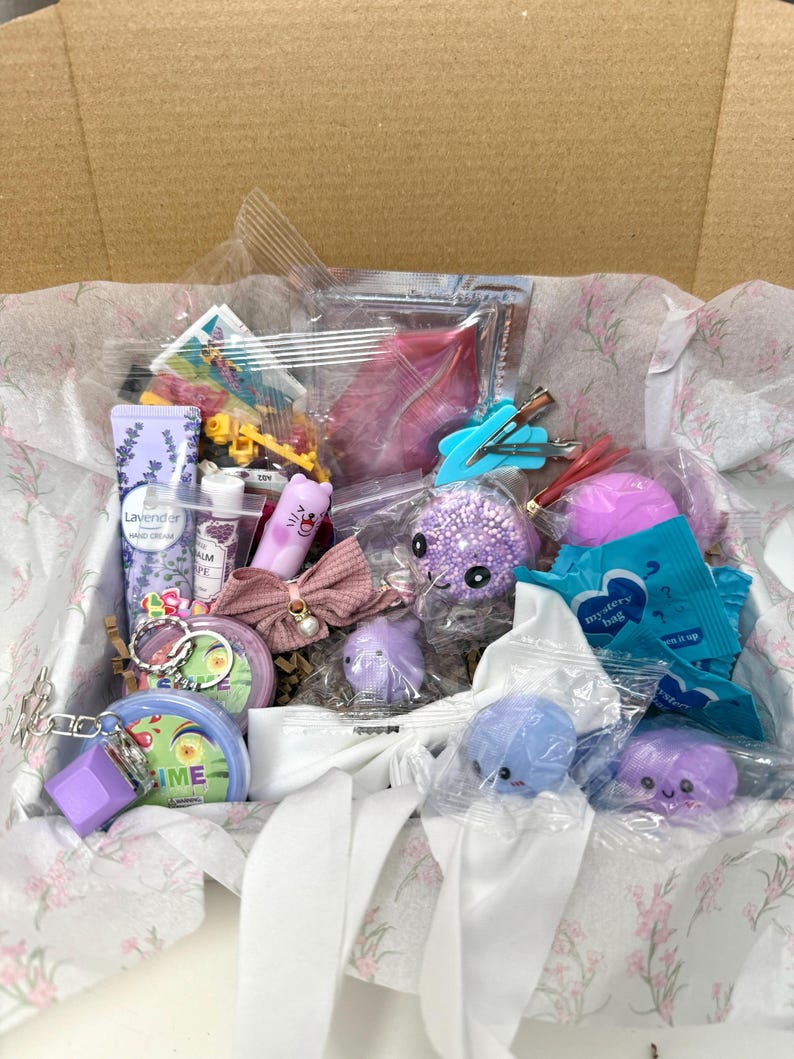 Puede incluir: Una caja de regalo llena de art&iacute;culos peque&ntilde;os variados, incluyendo un tubo de crema de manos de lavanda, un lazo de tela rosa con un detalle de perla, varias figuras de animales blandas moradas y azules, una bolsa misteriosa azul, slime y horquillas.