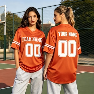 May include: An orange jersey with white V-neck trim and white stripes on the sleeves. The front features the text TEAM NAME above the number 00 in white. The back displays the text YOUR NAME above the number 00 in white. Paired with gray pants.