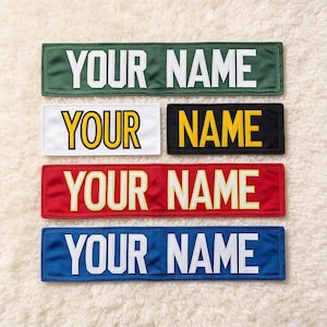 May include: Five rectangular fabric patches with stitched borders. The top patch is green with white text, the middle left is white with yellow text, the middle right is black with yellow text, the red has yellow text, and the bottom is blue with white text. All read YOUR NAME.