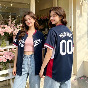 May include: A navy blue baseball jersey with white and red trim, featuring the text Korea on the front and the text Your Name with the number 00 on the back. The jersey is worn open over a light pink crop top and light blue denim jeans.