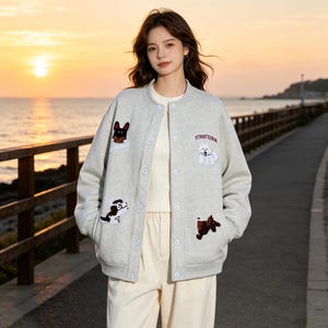 Puede incluir: Una chaqueta bomber gris claro abotonada con parches bordados de perros y el texto STARFEINK. La chaqueta tiene cuello acanalado, mangas largas y un corte relajado, usada sobre una prenda superior blanca lisa y pantalones claros.
