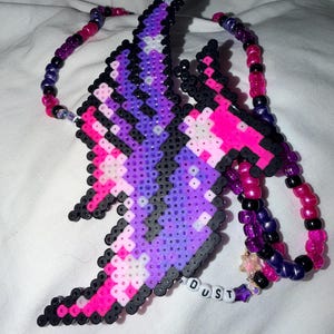 May include: A necklace featuring a large, pixelated pendant made of fused plastic beads in shades of purple, pink, black, and white. The pendant is attached to a beaded chain of alternating pink, purple, and black beads, with white letter beads spelling DUST.