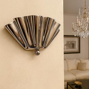 Könnte beinhalten: Eine Wandleuchte mit poliertem Chrom-Finish und einem fächerförmigen, plissierten Muscheldesign. Die Leuchte hat eine halbkreisförmige Silhouette mit vertikalen Rillen, die von einem zentralen kugelförmigen Akzent an der Basis ausgehen.