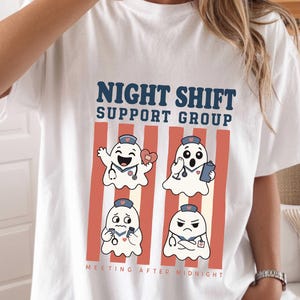 Può includere: Una t-shirt bianca con il testo NIGHT SHIFT SUPPORT GROUP e MEETING AFTER MIDNIGHT. La grafica mostra quattro fantasmi dei cartoni animati che indossano cuffie da infermiere e stetoscopi, con espressioni diverse, su strisce verticali rosse.