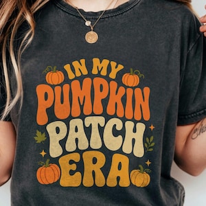 Può includere: Una t-shirt a maniche corte grigio antracite con una stampa grafica che riporta il testo "IN MY PUMPKIN PATCH ERA" in caratteri rétro arancioni e crema, arricchita da illustrazioni di zucche arancioni, piccole foglie verdi e stelle.