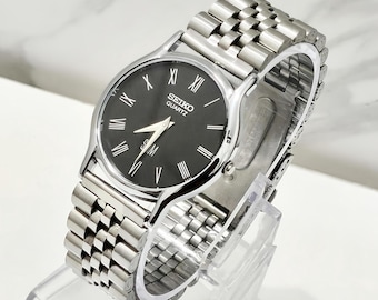 Könnte beinhalten: Eine silberfarbene Seiko Quartz Armbanduhr mit einem runden schwarzen Zifferblatt und weißen römischen Ziffern als Stundenmarkierungen. Die Uhr verfügt über ein mehrgliedriges Edelstahlarmband und silberne Zeiger. Seiko und Quartz sind aufgedruckt.