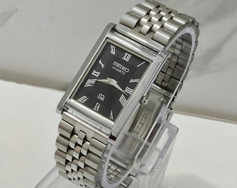 Könnte beinhalten: Eine rechteckige Seiko Quartz Armbanduhr mit schwarzem Zifferblatt, silberfarbenen römischen Ziffern als Stundenmarkierungen und silbernen Zeigern. Die Uhr verfügt über ein poliertes Edelstahlgehäuse und ein mehrgliedriges Metallarmband.