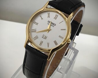 Könnte beinhalten: Eine goldfarbene Seiko Quartz Slim Armbanduhr mit weißem Zifferblatt, schwarzen römischen Ziffern und goldfarbenen Zeigern. Die Uhr verfügt über ein schwarzes Lederarmband mit strukturierter Oberfläche und eine goldfarbene Krone am Gehäuse.