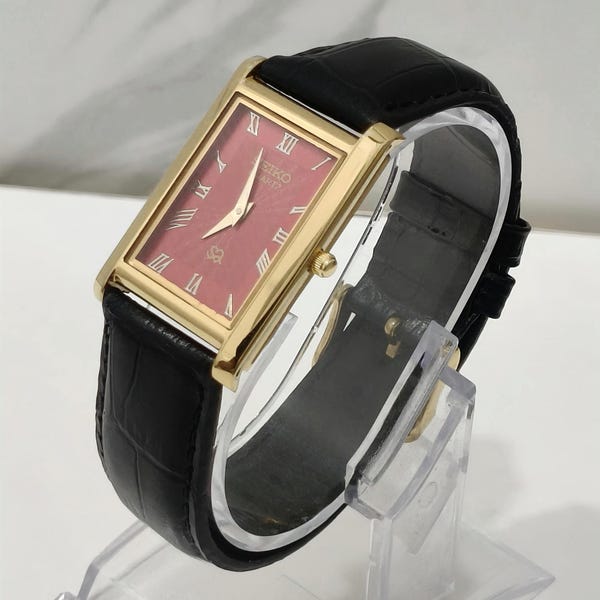 May include: A rectangular Seiko Quartz watch with a gold-tone case and a deep red dial. The dial features gold-tone Roman numeral hour markers and gold-tone hands. The watch is fitted with a black leather strap that has a textured crocodile pattern.