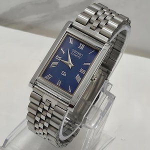 May include: A rectangular Seiko Quartz wristwatch with a deep blue dial, silver-tone Roman numeral hour markers, and silver hands. The watch features a polished silver-tone metal link bracelet and a small crown on the right side of the case.