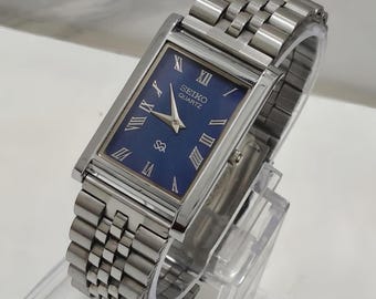 Könnte beinhalten: Eine rechteckige Seiko Quartz Armbanduhr mit tiefblauem Zifferblatt, silberfarbenen römischen Ziffern als Stundenmarkierungen und silbernen Zeigern. Die Uhr hat ein poliertes silberfarbenes Metallgliederarmband und eine kleine Krone.