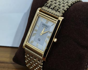 Könnte beinhalten: Eine rechteckige, goldfarbene Seiko Quartz Armbanduhr mit hellem Zifferblatt mit Wellenmuster, goldenen römischen Ziffern als Stundenmarkierungen und goldenen Zeigern. Die Uhr hat ein passendes, goldfarbenes Metallgliederarmband mit Struktur.
