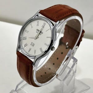 May include: A Seiko Quartz Slim wristwatch featuring a round silver-tone case, a white dial with black Roman numeral hour markers, and a brown textured leather strap with a buckle closure. The watch face displays the text SEIKO QUARTZ and SLIM.