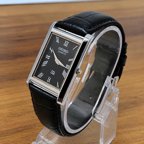 May include: A rectangular Seiko Quartz wristwatch featuring a black dial with silver-toned Roman numeral hour markers and hands. The watch has a polished silver-tone metal case and a black leather strap with a textured crocodile-style pattern.