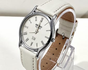 Könnte beinhalten: Eine Seiko Quartz Slim Armbanduhr mit rundem silberfarbenem Gehäuse, weißem Zifferblatt mit schwarzen römischen Ziffern und einem weißen Lederarmband. Auf dem Zifferblatt stehen die Wörter Seiko, Quartz und Slim in schwarzer Schrift.