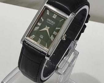 Könnte beinhalten: Eine rechteckige Seiko Quartz Armbanduhr mit dunkelgrünem Zifferblatt, silberfarbenen römischen Ziffern als Stundenmarkierungen und silbernen Zeigern. Die Uhr hat ein poliertes silberfarbenes Metallgehäuse und ein schwarzes Lederarmband.