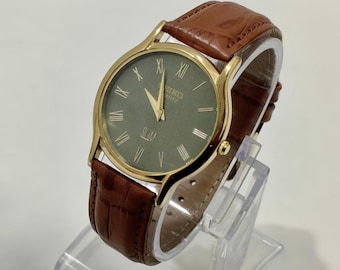 Könnte beinhalten: Eine Seiko Quartz Slim Armbanduhr mit einem runden goldfarbenen Gehäuse, einem dunkelolivgrünen Zifferblatt mit goldfarbenen römischen Ziffern und goldfarbenen Zeigern. Die Uhr hat ein braunes Lederarmband mit einer strukturierten Krokodilprägung.