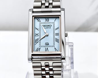Könnte beinhalten: Eine rechteckige Seiko Quartz Armbanduhr mit hellblauem Zifferblatt und schwarzen römischen Ziffern. Die Uhr verfügt über ein poliertes silberfarbenes Edelstahlgehäuse und ein passendes Gliederarmband aus Metall mit Sicherheitsverschluss.