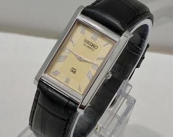 Könnte beinhalten: Eine rechteckige Seiko Quartz Armbanduhr mit cremefarbenem Zifferblatt, silberfarbenen römischen Ziffern als Stundenmarkierungen und Zeigern. Die Uhr hat ein poliertes silberfarbenes Metallgehäuse und ein schwarzes Lederarmband mit Krokoprägung.