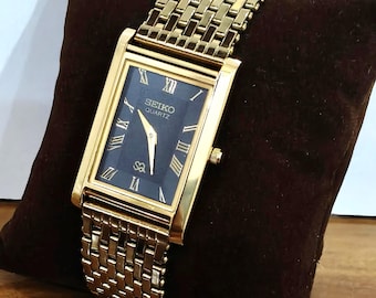 Könnte beinhalten: Eine rechteckige, goldfarbene Seiko Quartz Armbanduhr mit dunkelblauem Zifferblatt, goldenen römischen Ziffern als Stundenmarkierungen, goldenen Zeigern und einem passenden, strukturierten Gliederarmband aus goldfarbenem Metall.