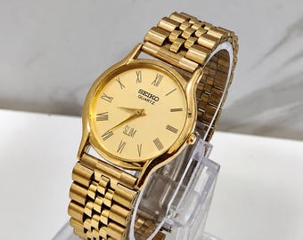 Könnte beinhalten: Eine goldfarbene Seiko Quartz Armbanduhr mit rundem Gehäuse, goldenem Zifferblatt und römischen Ziffern. Die Uhr hat ein flaches Profil, goldene Zeiger und ein passendes goldfarbenes Metallgliederarmband mit feinem Gliederdesign.