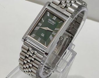 Könnte beinhalten: Eine rechteckige Seiko Quartz Armbanduhr mit einem dunkelgrünen, strukturierten Zifferblatt, silberfarbenen römischen Ziffern als Stundenmarkierungen und silbernen Zeigern. Die Uhr hat ein poliertes Edelstahlgehäuse und ein passendes Gliederarmband.