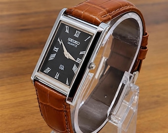 Könnte beinhalten: Eine rechteckige Seiko Quartz Armbanduhr mit schwarzem Zifferblatt, silberfarbenen römischen Ziffern als Stundenmarkierungen und goldfarbenen Zeigern. Die Uhr hat ein poliertes silberfarbenes Metallgehäuse und ein strukturiertes braunes Lederarmband.