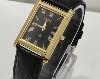 Könnte beinhalten: Eine rechteckige Seiko Quartz Armbanduhr mit goldfarbenem Gehäuse und schwarzem Zifferblatt mit goldenen römischen Ziffern. Die Uhr verfügt über goldfarbene Zeiger und ein schwarzes Lederarmband mit einer strukturierten Krokodilprägung.