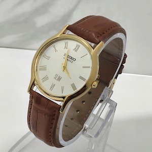 Op de afbeelding: Een goudkleurig polshorloge met een witte wijzerplaat, gouden wijzers en zwarte Romeinse cijfers. Op de wijzerplaat staat de tekst SEIKO QUARTZ en SLIM. Het horloge heeft een bruine lederen band met een gestructureerd, krokodilachtig reliëf.