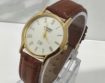 Könnte beinhalten: Eine goldfarbene Armbanduhr mit weißem Zifferblatt, goldenen Zeigern und schwarzen römischen Ziffern. Auf dem Zifferblatt stehen die Schriftzüge SEIKO QUARTZ und SLIM. Sie hat ein braunes Lederarmband mit einer strukturierten Krokodilprägung.
