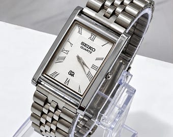 Könnte beinhalten: Eine rechteckige Seiko Quartz Armbanduhr mit einem silberfarbenen Edelstahl-Gliederarmband und einem weißen Zifferblatt. Das Zifferblatt zeigt schwarze römische Ziffern, silberne Zeiger und den Seiko Quartz Schriftzug in Schwarz.
