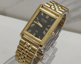 Könnte beinhalten: Eine rechteckige, goldfarbene Armbanduhr mit schwarzem Zifferblatt, goldenen römischen Ziffern und goldenen Zeigern. Die Uhr verfügt über ein passendes, goldfarbenes Metallgliederarmband und trägt den Schriftzug SEIKO QUARTZ.