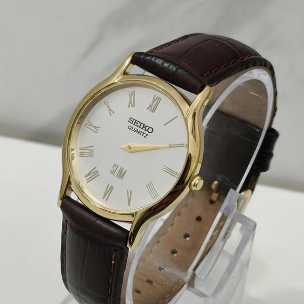 May include: A Seiko Quartz Slim wristwatch featuring a round gold-tone case, a white dial with black Roman numeral hour markers, and gold-tone hands. The watch is fitted with a dark brown leather strap that has a textured, embossed finish.