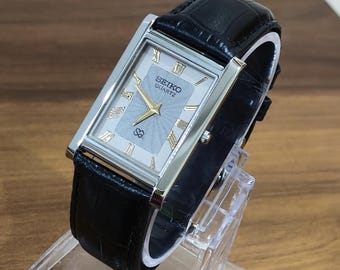 Könnte beinhalten: Eine rechteckige Seiko Quartz Armbanduhr mit silberfarbenem Zifferblatt mit strukturiertem Strahlenmuster, goldfarbenen Zeigern und goldenen römischen Ziffern. Die Uhr hat ein poliertes silberfarbenes Gehäuse und ein schwarzes Lederarmband.