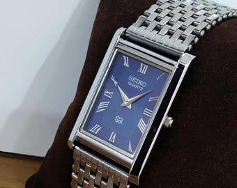 Könnte beinhalten: Eine rechteckige Seiko Quartz Armbanduhr mit tiefblauem Zifferblatt, silberfarbenen römischen Ziffern als Stundenmarkierungen und silbernen Zeigern. Die Uhr hat ein poliertes silberfarbenes Metallgehäuse und ein passendes Gliederarmband.